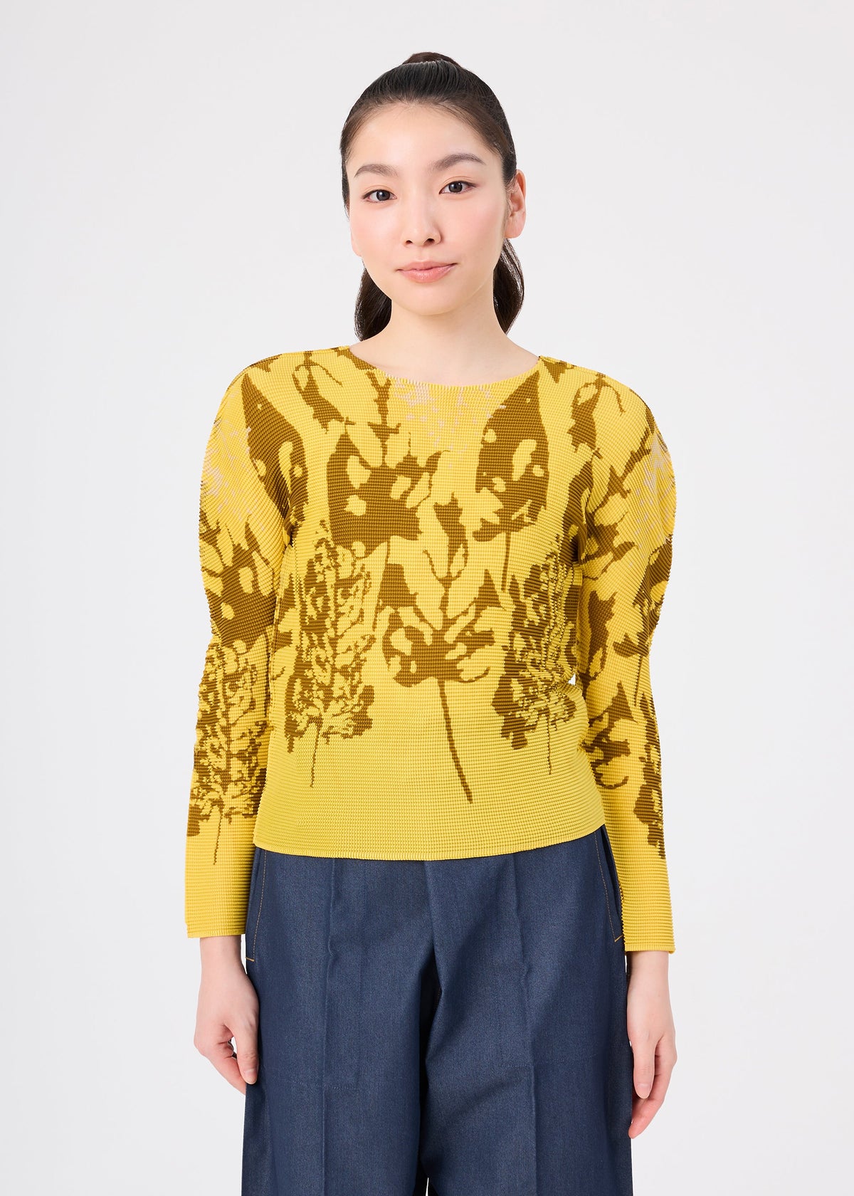 Zoomed view A woman with long dark hair in a ponytail wears the LEAF top by me ISSEY MIYAKE, featuring yellow fabric with brown leaf patterns, paired with dark blue wide-leg pants against a white background.