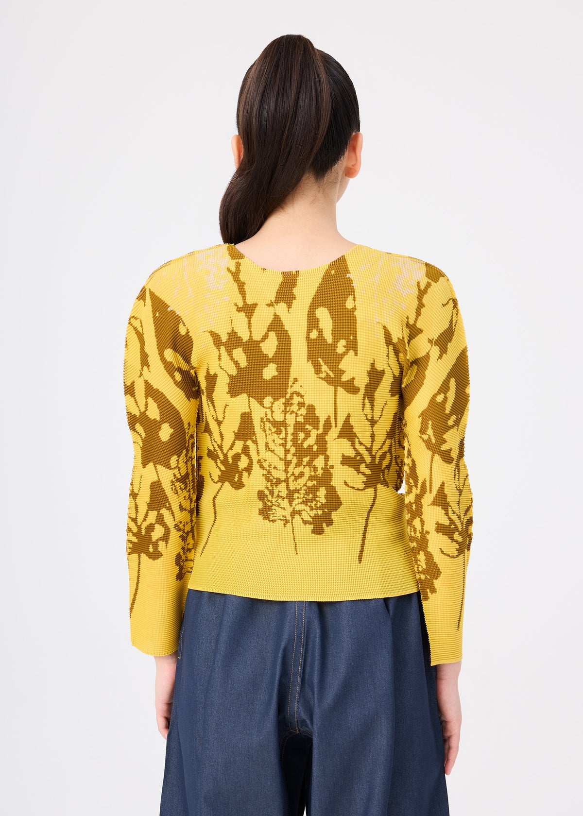 A person with a sleek ponytail faces away, wearing the LEAF yellow round-neck top with brown abstract leaf pattern by me ISSEY MIYAKE and blue high-waisted pants, set against a plain white background. view 6 of 7