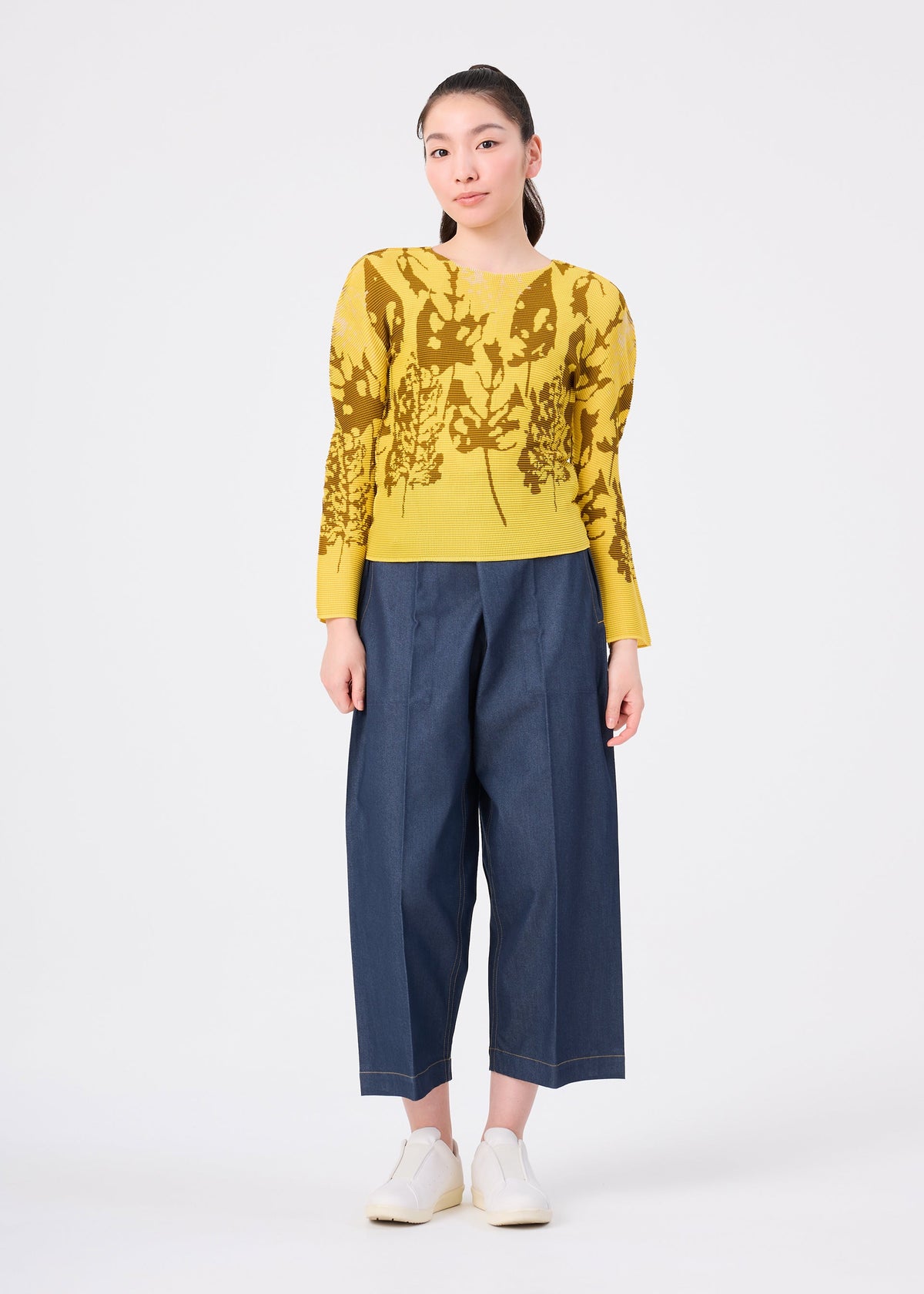 Zoomed view A person wears the LEAF top by me ISSEY MIYAKE—yellow, round-neck, pleated with a leaf pattern—paired with wide-leg blue pants and white slip-on shoes, standing against a white background.