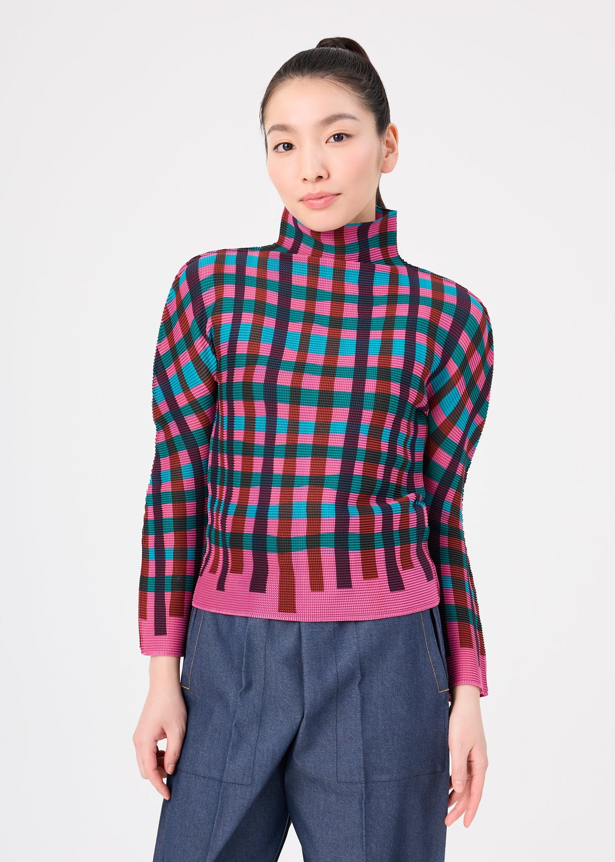 Zoomed view A woman with long dark hair in a ponytail wears the TAPE CHECK top by me ISSEY MIYAKE and blue high-waisted pants, standing against a plain white background.