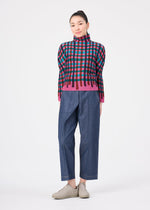 Woman standing against a white background, wearing the TAPE CHECK top by me ISSEY MIYAKE with wide-leg blue jeans and beige slip-on shoes. Her hair is tied back as she looks at the camera with a slight smile.