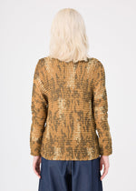 A person with wavy, shoulder-length blonde hair wears the LEAF PLEATS SHIRT by me ISSEY MIYAKE in gold and brown, styled with dark pants, shown from the back against a plain white background.