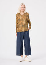 A person with light hair wears the LEAF PLEATS SHIRT by me ISSEY MIYAKE, wide-leg blue pants, and white shoes, standing against a white background with a slight smile and one hand in their pocket.