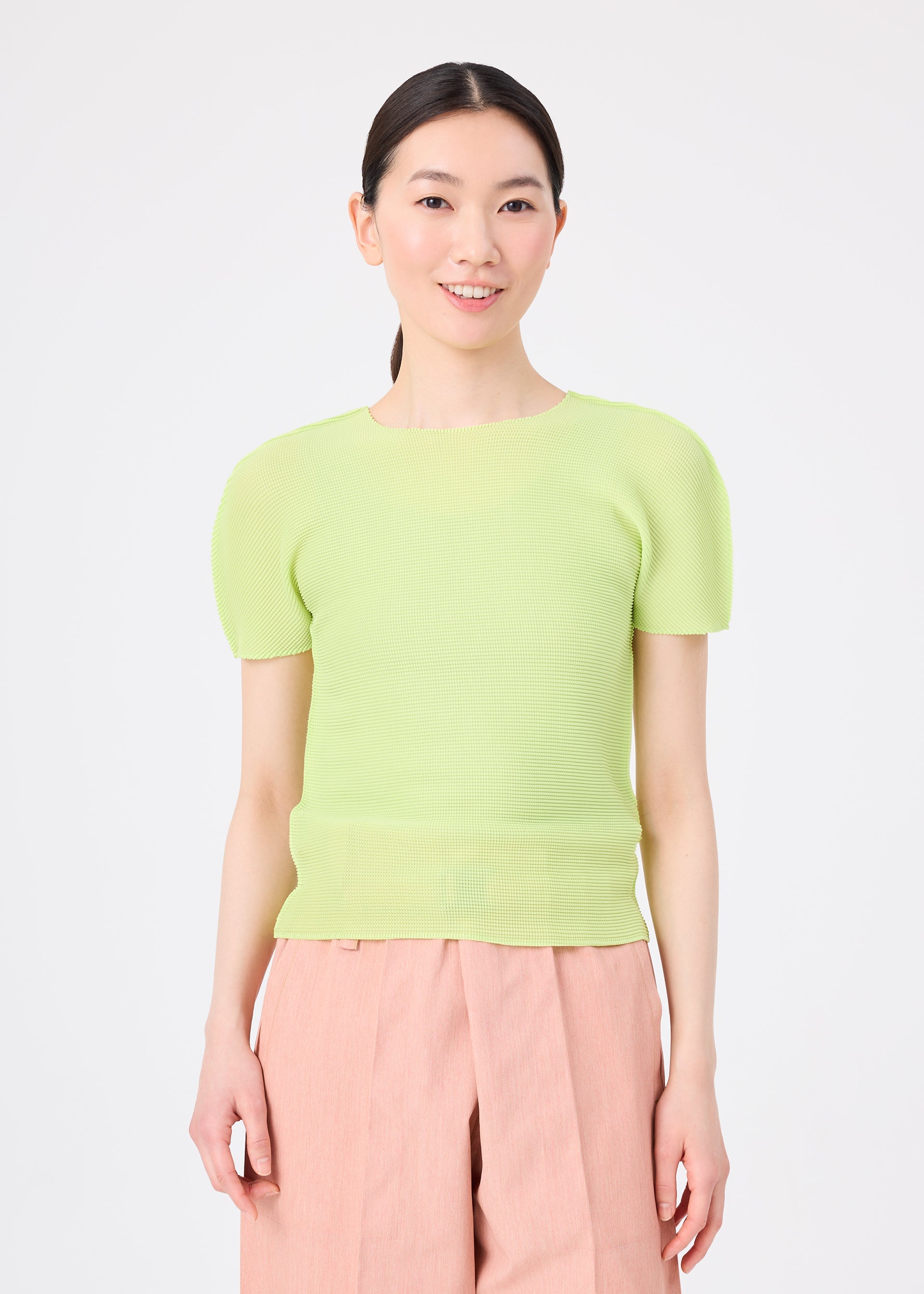 Zoomed view A person with long dark hair tied back is wearing the me ISSEY MIYAKE STRETCH PLEATS 1 top in light green and high-waisted light pink pants, smiling against a plain white background.