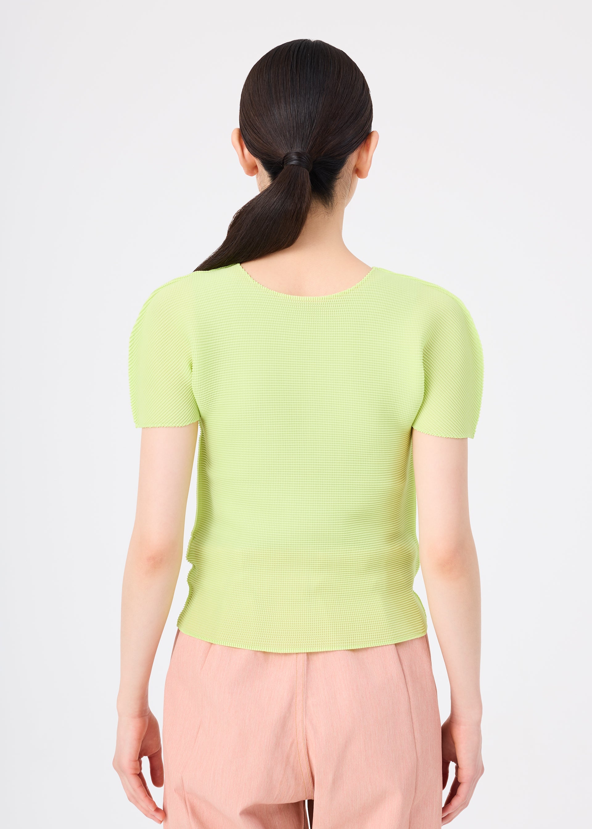 Zoomed view A person with long dark hair in a low ponytail is seen from behind, wearing the me ISSEY MIYAKE STRETCH PLEATS 1 light green short-sleeved top and light pink pants against a plain white background.