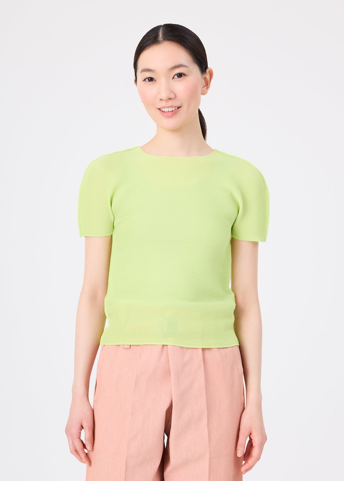 A woman with long dark hair tied back wears the STRETCH PLEATS 1 by me ISSEY MIYAKE—a light green round-neck short-sleeved top and high-waisted light pink trousers—standing and smiling against a plain white background. view 8 of 8