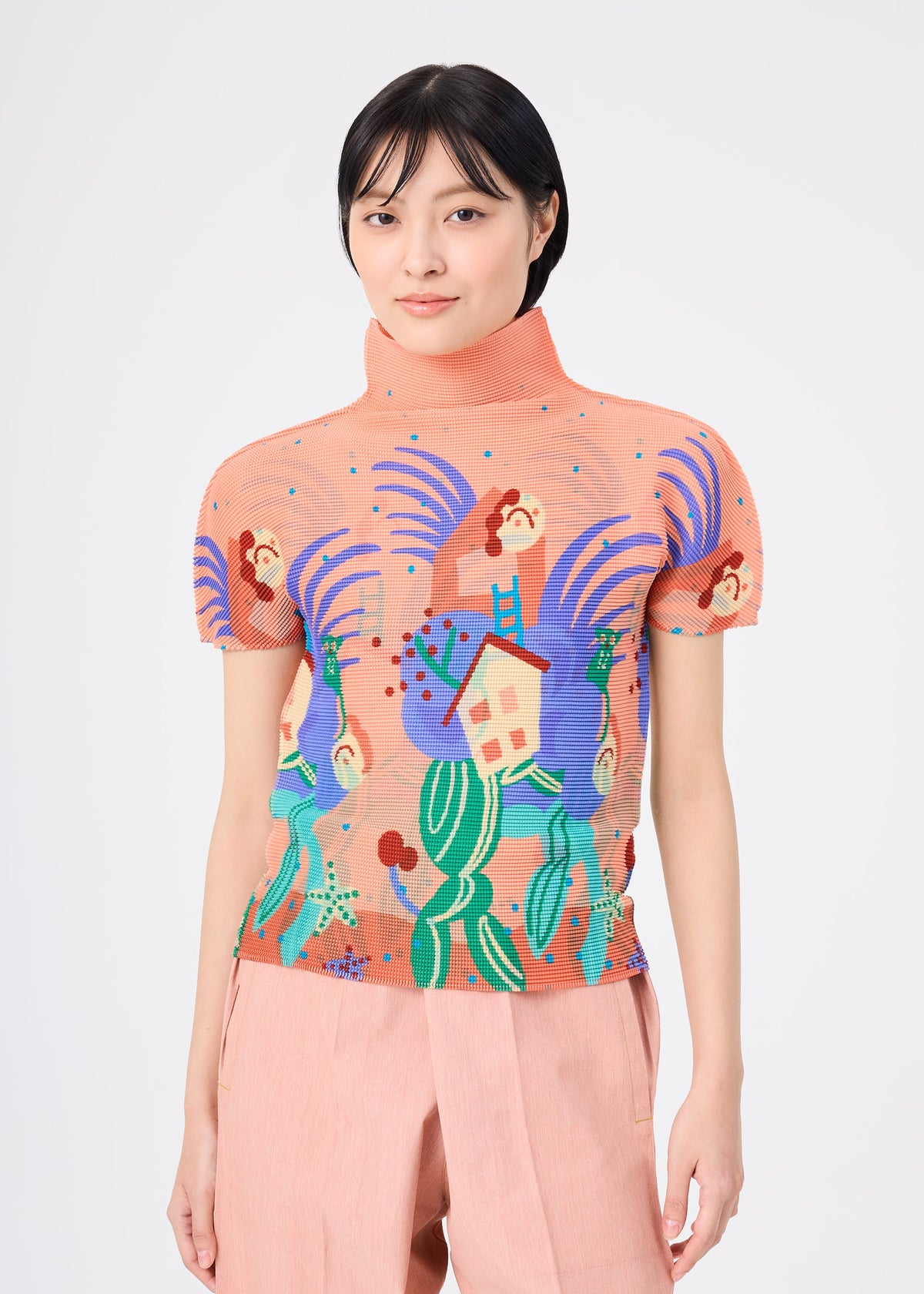 A woman with short black hair wears the ISLAND top by me ISSEY MIYAKE, a peach-colored stretch pleats piece with a vibrant abstract print, paired with light peach pants against a plain background. view 4 of 6