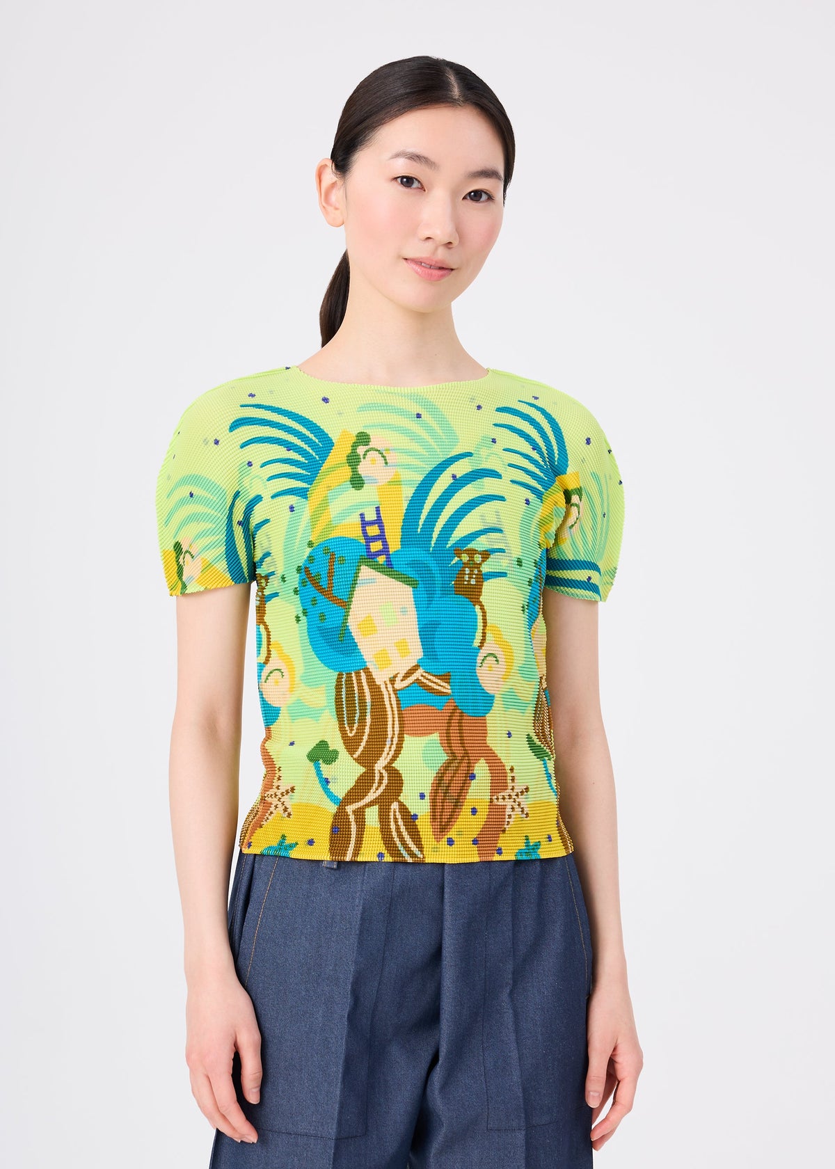 A woman with long dark hair in a low ponytail wears the ISLAND short-sleeved stretch pleats top by me ISSEY MIYAKE, featuring colorful Philippine motifs, paired with blue high-waisted pants against a plain light background. view 4 of 6