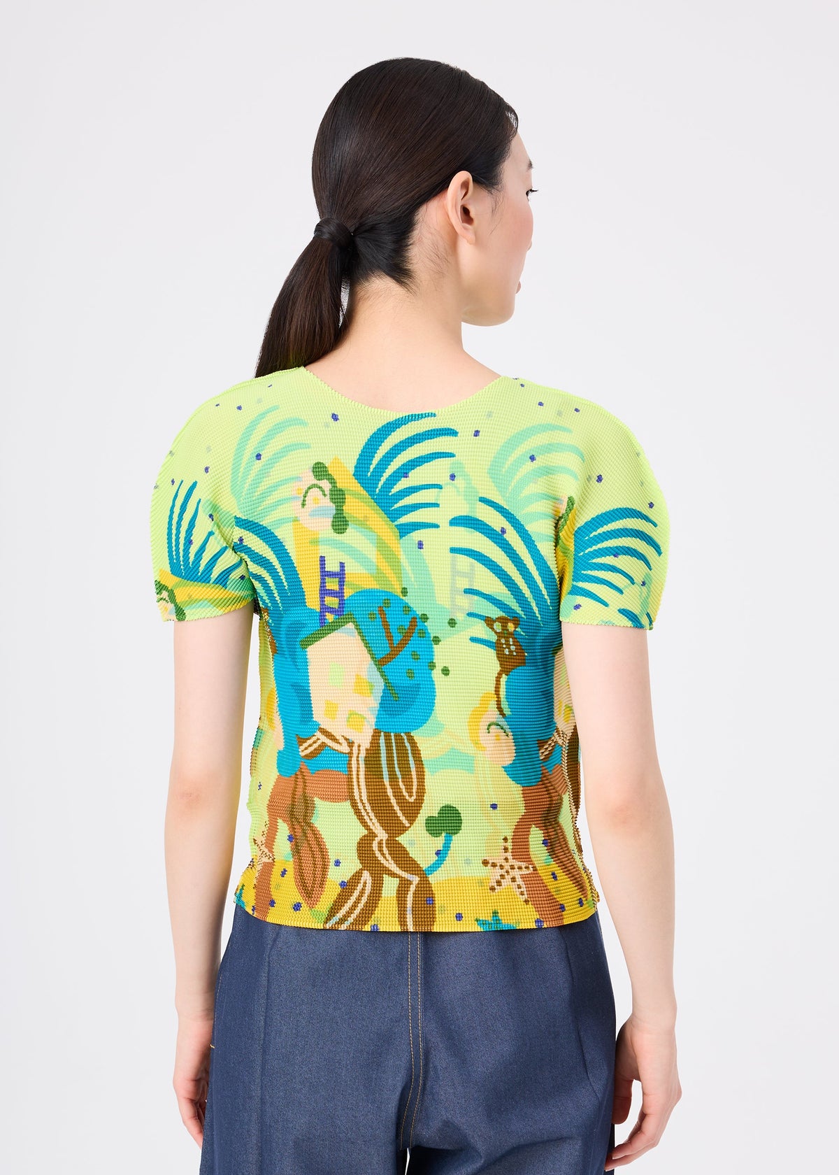 A woman with dark hair in a low ponytail is seen from the back, wearing the ISLAND stretch pleats top by me ISSEY MIYAKE, featuring abstract blue, green, and brown Philippine-inspired patterns with dark blue pants against a plain background. view 5 of 6
