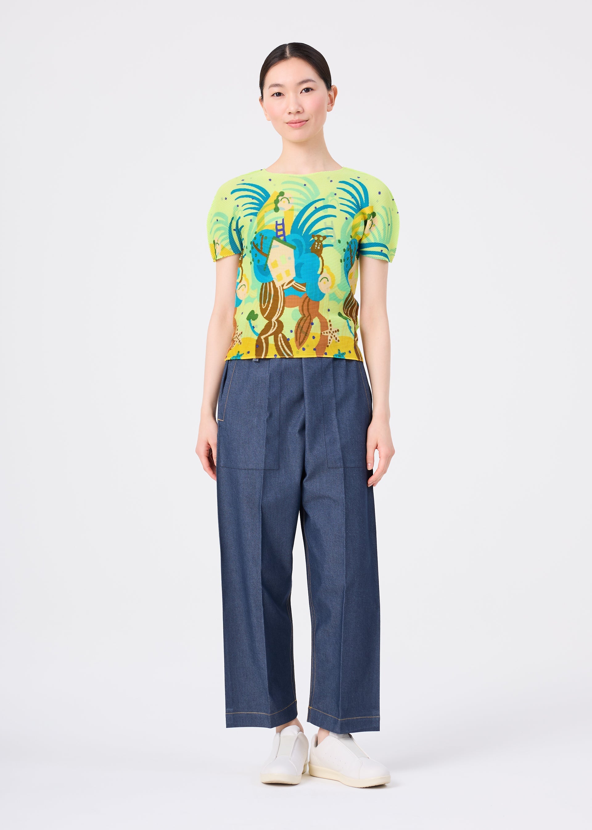 Zoomed view A woman faces forward in a light green ISLAND stretch pleats top with a colorful tarsier pattern by me ISSEY MIYAKE, paired with loose blue denim pants and white shoes against a plain white background.