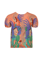 The ISLAND shirt by me ISSEY MIYAKE is a short-sleeved orange piece showcasing stylized roosters, houses, and green foliage in bold purple, red, and green hues inspired by Philippine motifs.