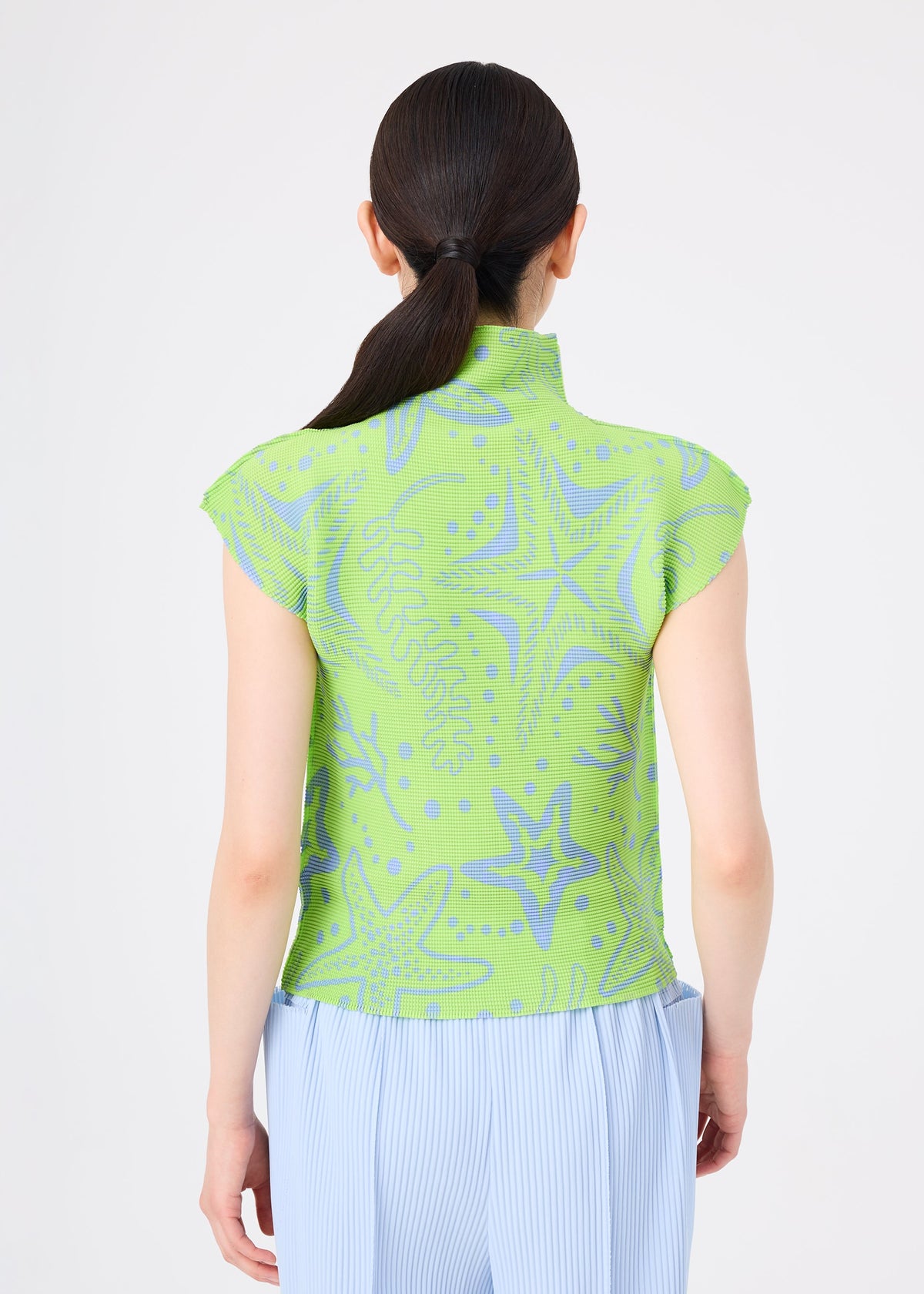 A woman with long dark hair in a low ponytail is seen from the back wearing the STARFISH top and light blue stretch pleat pants by me ISSEY MIYAKE, set against a plain white background. view 6 of 7
