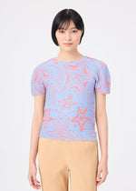 A woman with short black hair wears the me ISSEY MIYAKE STARFISH light blue knit top with a starfish and seaweed pattern, paired with beige pleated pants, standing against a plain white background.