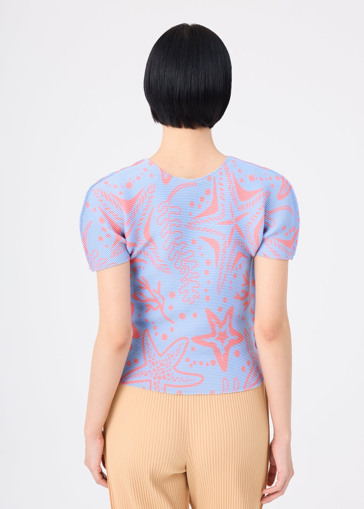 A person with short black hair is seen from behind wearing the me ISSEY MIYAKE STARFISH light blue round-neck short-sleeved shirt with a pink starfish and seaweed pattern, paired with beige pleated pants against a plain white background. view 6 of 7