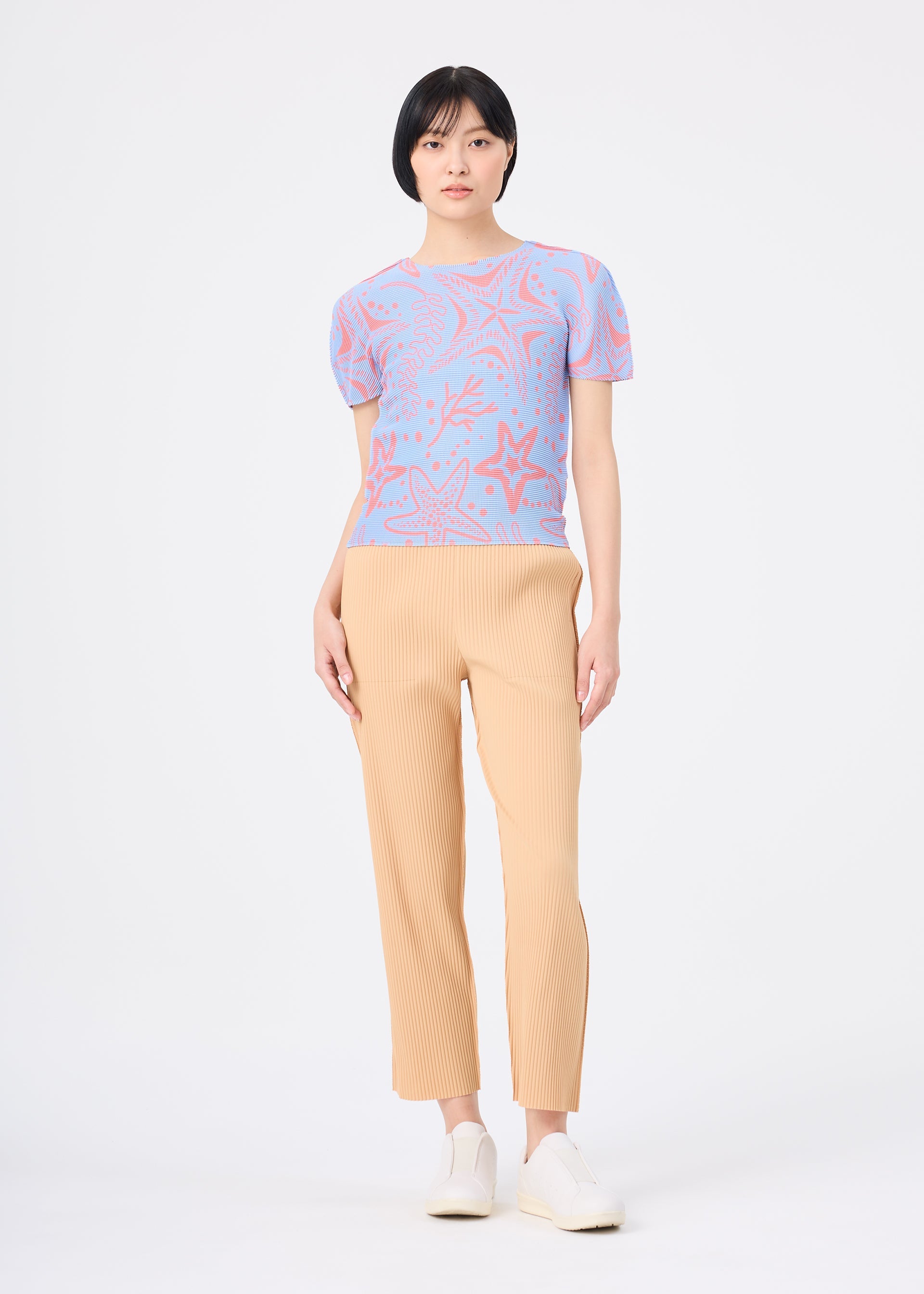 Zoomed view A woman faces forward against a white background, wearing the STARFISH short-sleeved, round-neck top with blue and pink starfish seaweed print by me ISSEY MIYAKE, beige pleated pants, and white shoes.