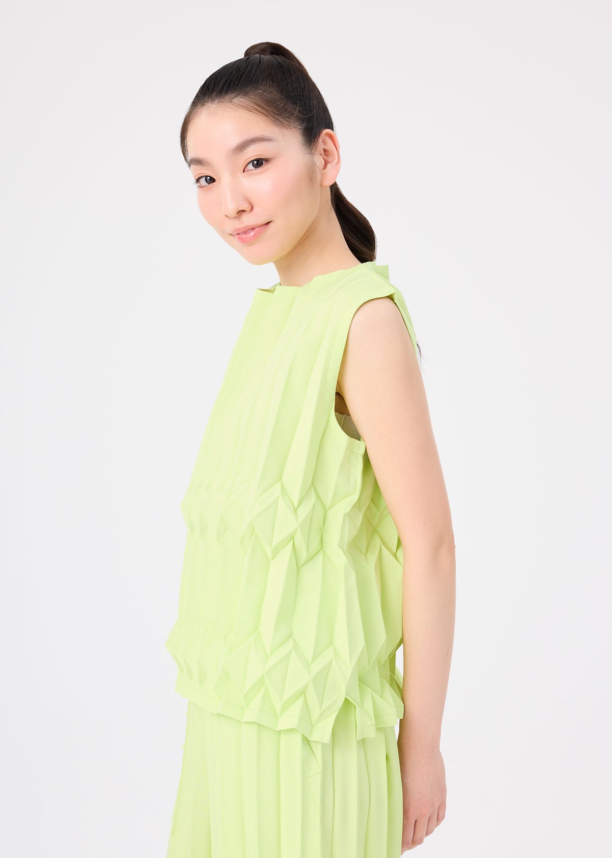 A young woman with long dark hair in a high ponytail smiles slightly, wearing the light green TREE PLEATS sleeveless top and skirt by me ISSEY MIYAKE, featuring a subtle three-dimensional pattern against a plain white background. view 8 of 8