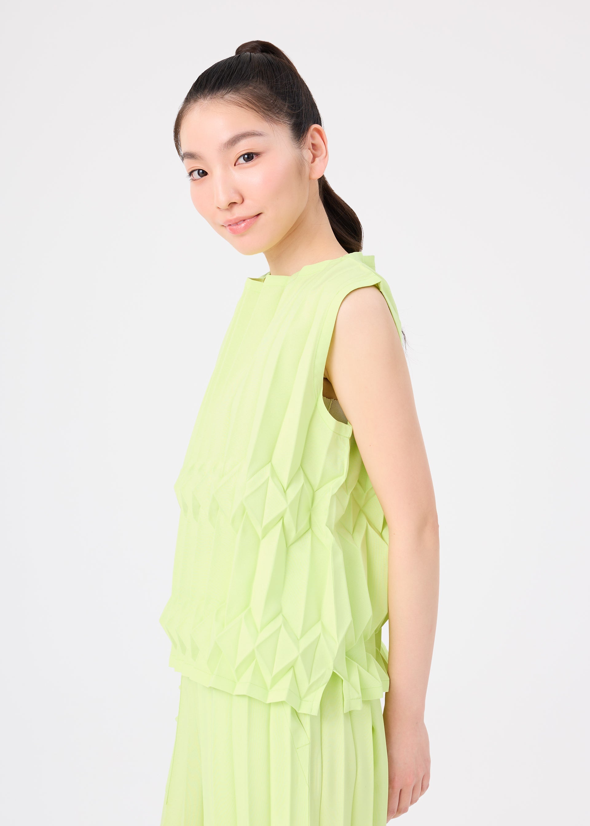 Zoomed view A young woman with long dark hair in a high ponytail smiles slightly, wearing the light green TREE PLEATS sleeveless top and skirt by me ISSEY MIYAKE, featuring a subtle three-dimensional pattern against a plain white background.