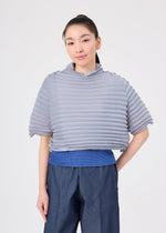 A person with long dark hair in a ponytail wears the me ISSEY MIYAKE ACCORDION STRETCH PLEATS top in light gray, featuring textured stretch pleats and a high neck. Paired with blue pants, they stand against a plain white background, smiling gently with one hand behind their back. The stretch pleats of the ACCORDION STRETCH PLEATS top enhance its comfortable feel.