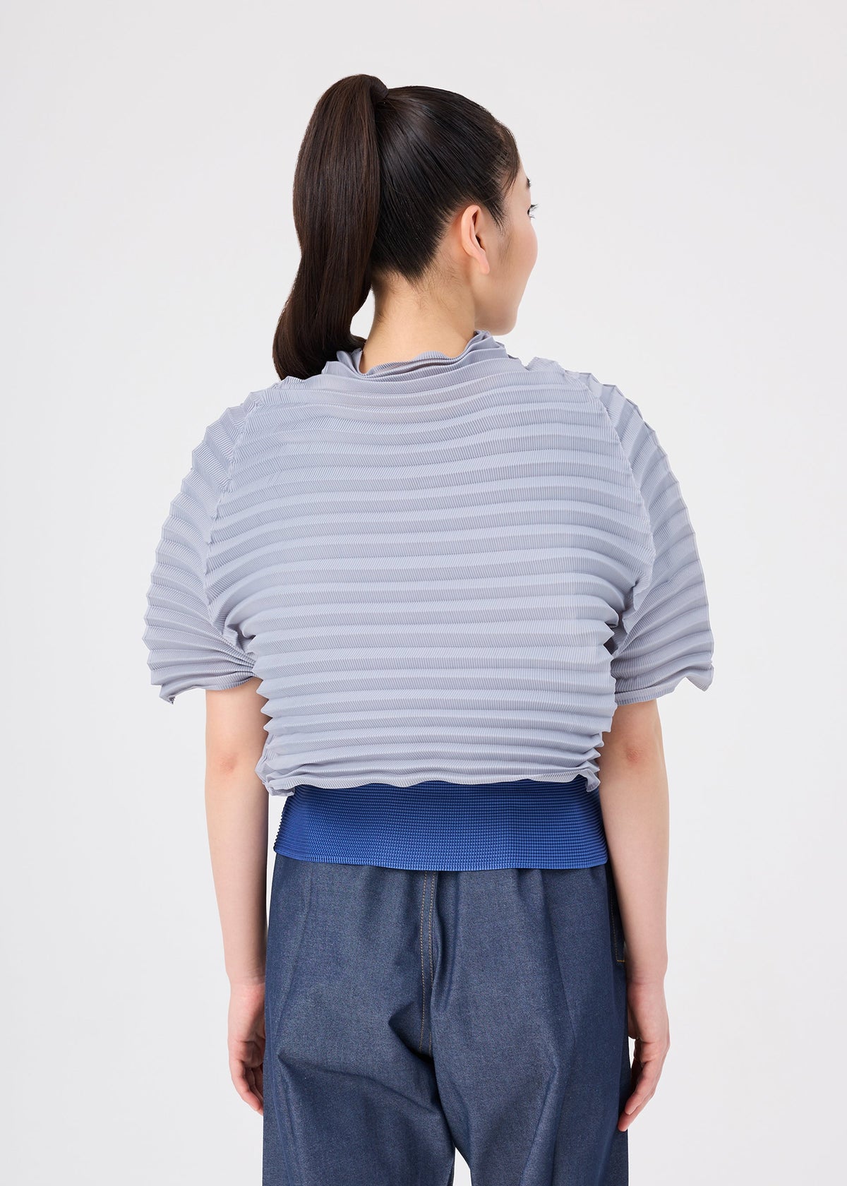 A person with a high ponytail is seen from behind, standing against a plain white background, wearing the me ISSEY MIYAKE ACCORDION STRETCH PLEATS ribbed light gray short-sleeve top layered over a blue shirt, paired with loose denim pants. view 6 of 7