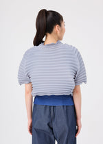 A person with a high ponytail is seen from behind, standing against a plain white background, wearing the me ISSEY MIYAKE ACCORDION STRETCH PLEATS ribbed light gray short-sleeve top layered over a blue shirt, paired with loose denim pants.