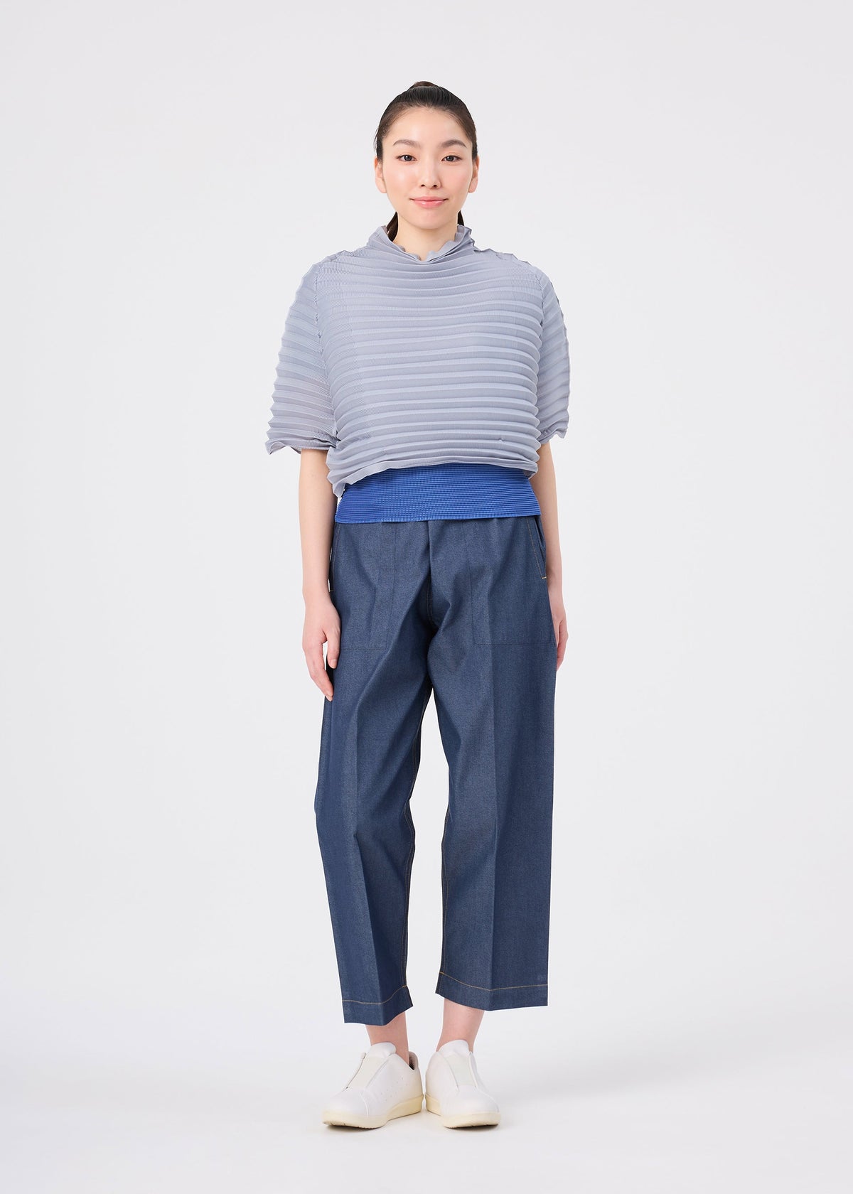 A person stands facing forward against a plain white background, dressed in me ISSEY MIYAKE’s ACCORDION STRETCH PLEATS: a textured light gray high-neck top layered over a blue shirt, with loose blue pants and white shoes. The outfit showcases the comfort and stylish appeal of stretch pleats. view 7 of 7