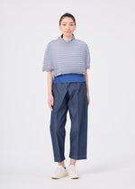 A person stands facing forward against a plain white background, dressed in me ISSEY MIYAKE’s ACCORDION STRETCH PLEATS: a textured light gray high-neck top layered over a blue shirt, with loose blue pants and white shoes. The outfit showcases the comfort and stylish appeal of stretch pleats.