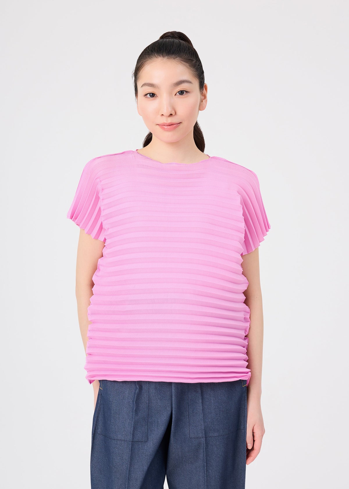 A person with long hair tied back wears the me ISSEY MIYAKE ACCORDION STRETCH PLEATS top in a textured, light pink round-neck short-sleeved style, paired with blue pants. They are standing against a plain white background and looking at the camera. view 5 of 7