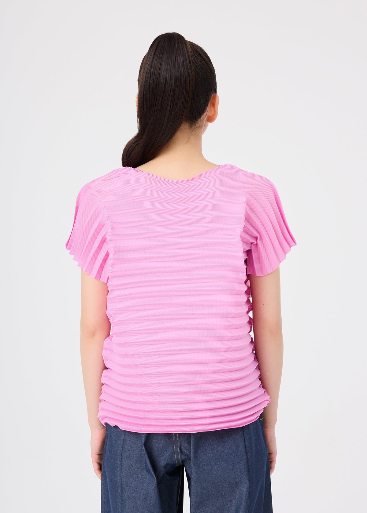 A person with long dark hair in a ponytail, wearing the me ISSEY MIYAKE ACCORDION STRETCH PLEATS blouse in pink with a round neck and short sleeves, paired with dark blue pants, stands with their back to the camera against a plain white background. view 6 of 7