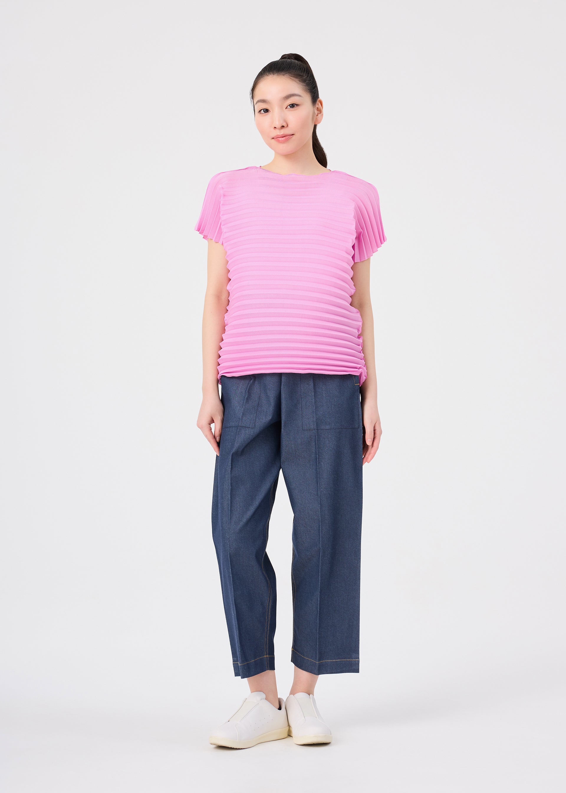 Zoomed view A woman stands facing forward against a plain white background, wearing the me ISSEY MIYAKE ACCORDION STRETCH PLEATS pink round-neck short-sleeved top, blue loose-fit pants, and white shoes.