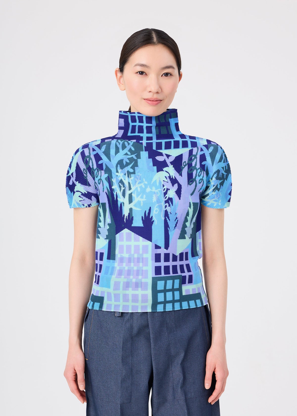 A person stands facing forward against a plain light background, wearing the BUILDING top by me ISSEY MIYAKE—a short-sleeve, high-neck design adorned with a blue, purple, and turquoise abstract forest pattern and delicate stretch pleats—paired with blue pants. view 5 of 7