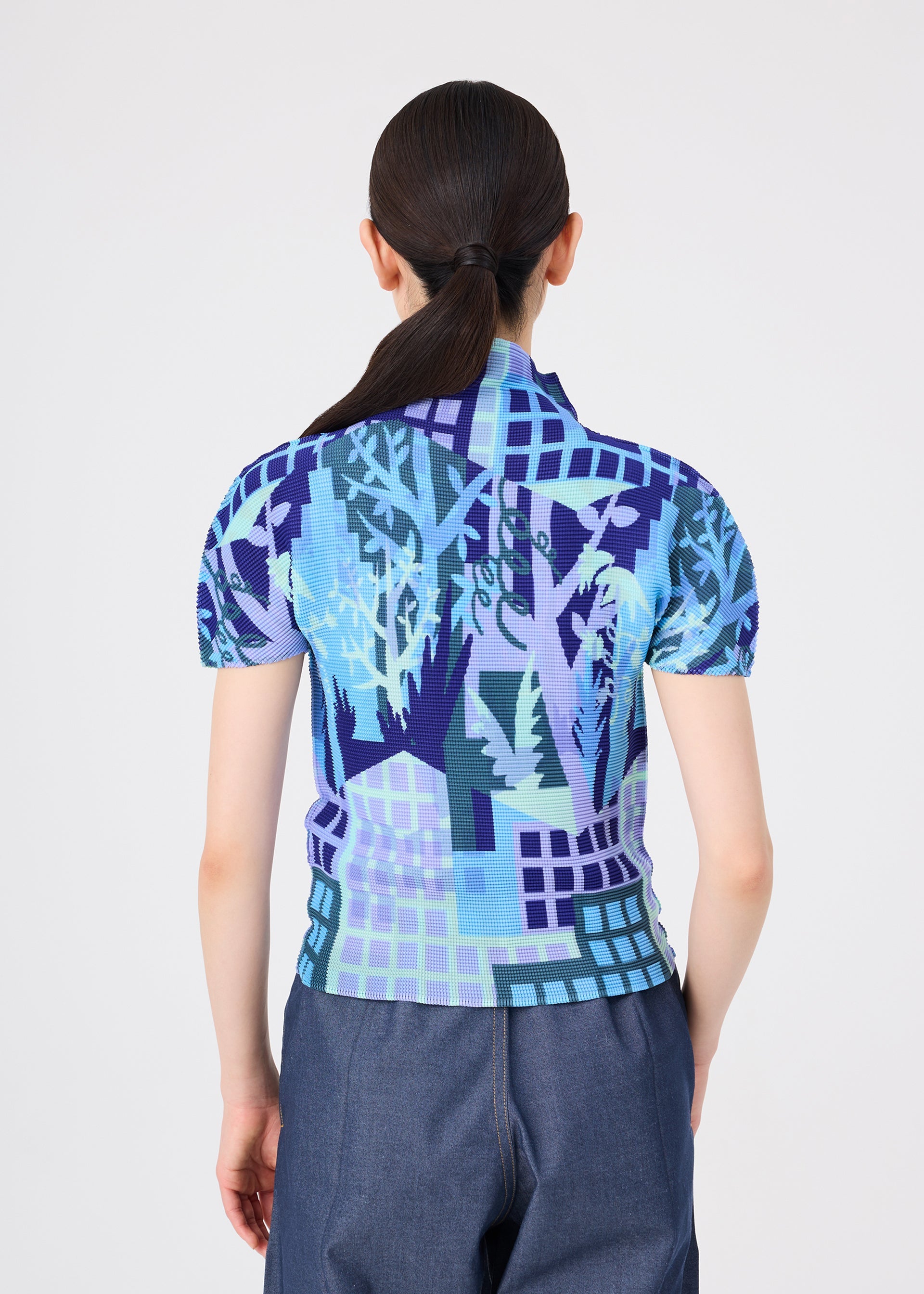 Zoomed view A person with long dark hair in a ponytail, wearing the short-sleeved blue patterned BUILDING shirt by me ISSEY MIYAKE—featuring motifs of trees and buildings inspired by urban farming scenes—stands facing away.