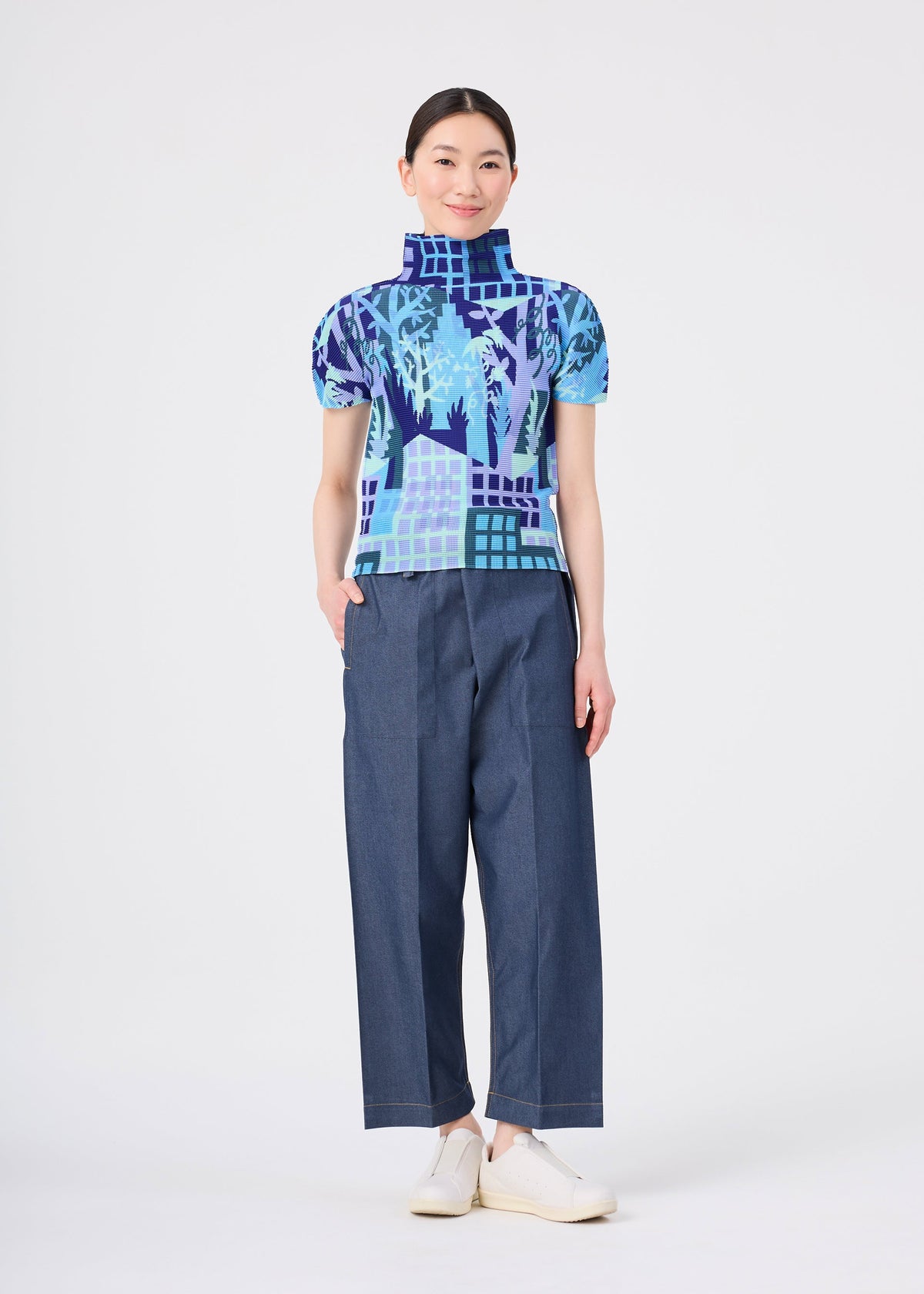 A person stands smiling against a plain white background, wearing the BUILDING blue patterned high-neck top with stretch pleats and wide-leg blue pants from me ISSEY MIYAKE, paired with white shoes. view 7 of 7