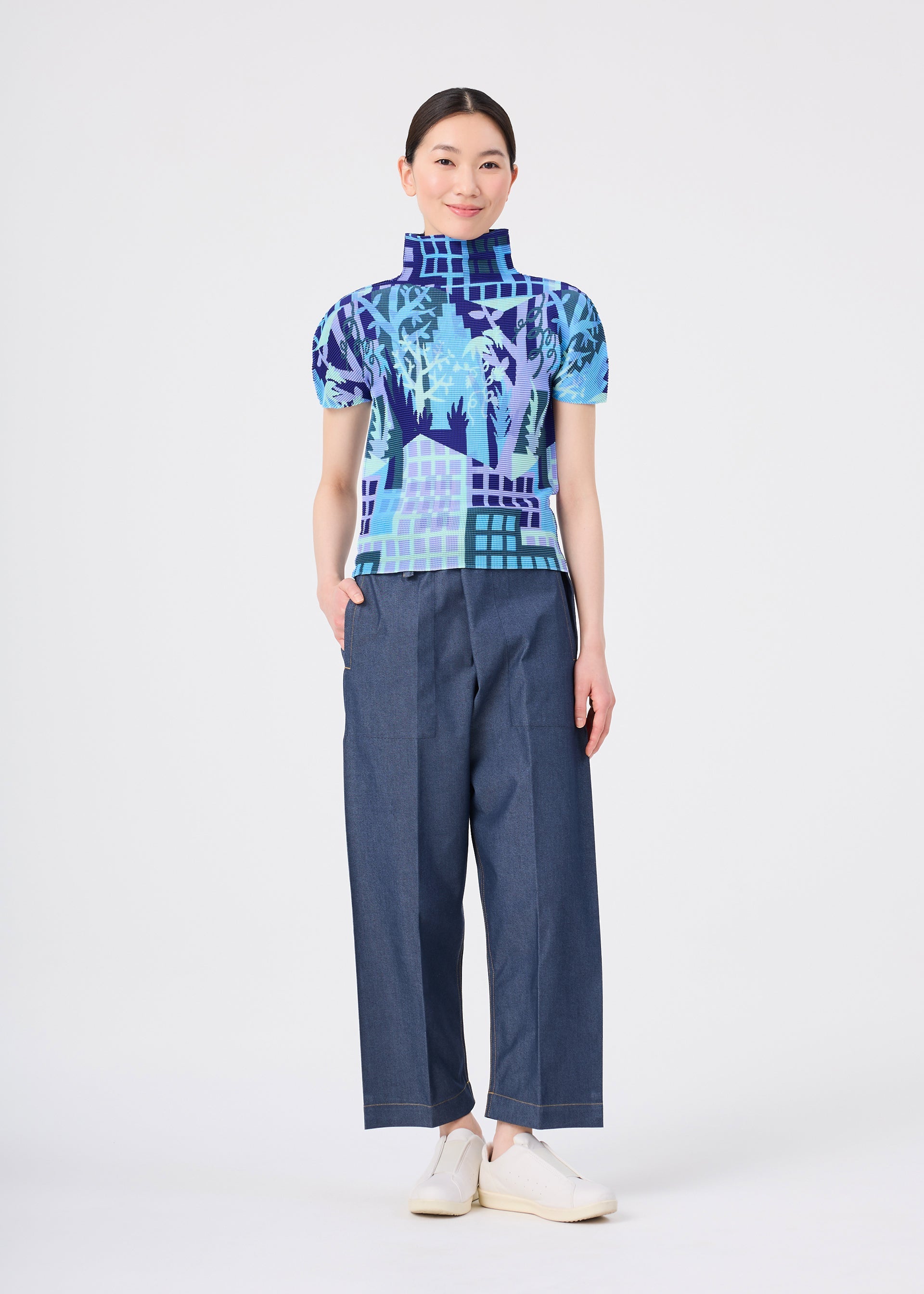 Zoomed view A person stands smiling against a plain white background, wearing the BUILDING blue patterned high-neck top with stretch pleats and wide-leg blue pants from me ISSEY MIYAKE, paired with white shoes.
