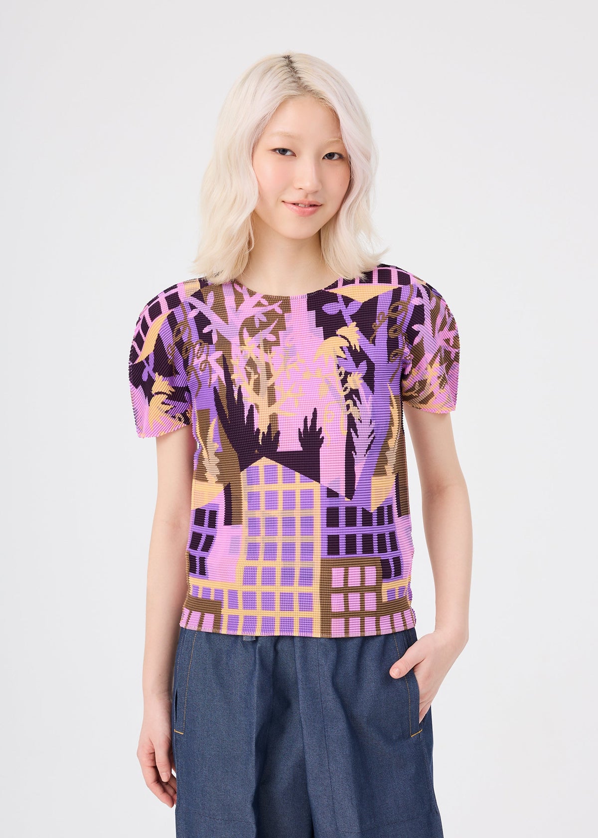 A smiling person with wavy, platinum blonde hair wears the BUILDING pleated round-neck top by me ISSEY MIYAKE, featuring a geometric palm tree pattern in purple, pink, and brown, paired with blue pants, standing against a plain white background. view 5 of 7