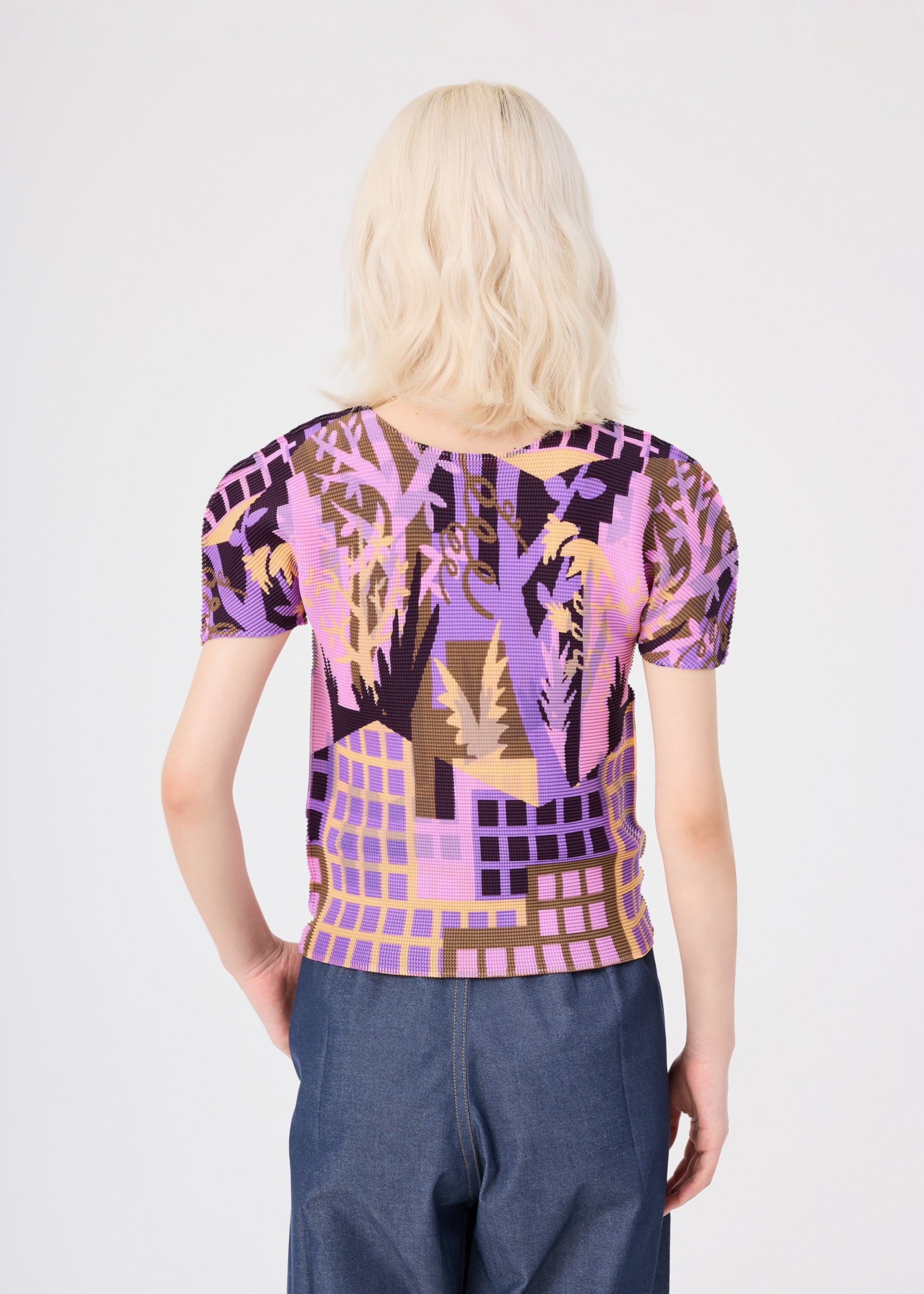 Zoomed view A person with wavy platinum blonde hair, seen from behind, is wearing the BUILDING short-sleeved pleated round-neck top by me ISSEY MIYAKE, which features a purple, pink, and brown abstract pattern, paired with blue high-waisted pants. The background is plain and light-colored.