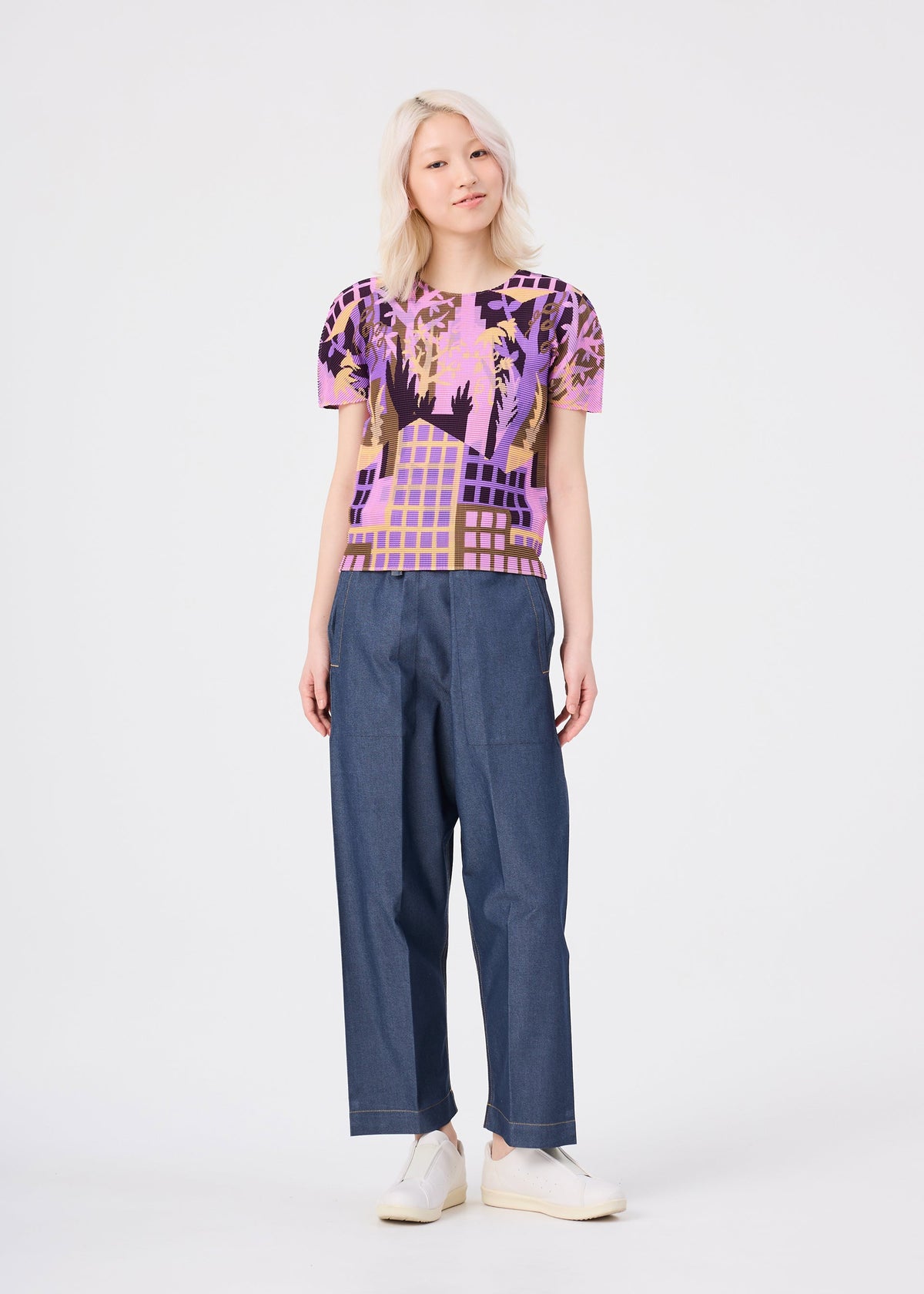 A person with light blonde hair wears the colorful “BUILDING” pleated round-neck top from me ISSEY MIYAKE, paired with loose blue pants and white slip-on shoes, standing against a plain white background. view 7 of 7