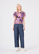 A person with light blonde hair wears the colorful “BUILDING” pleated round-neck top from me ISSEY MIYAKE, paired with loose blue pants and white slip-on shoes, standing against a plain white background.
