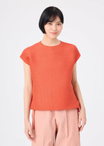 A person with short black hair is seen wearing the SQUARE STRETCH PLEATS top in orange from me ISSEY MIYAKE, along with light pink wide-leg pants, standing against a plain white background and smiling gently at the camera.