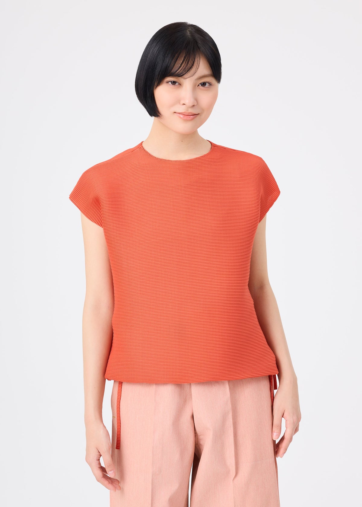 Zoomed view A person with short black hair is seen wearing the SQUARE STRETCH PLEATS top in orange from me ISSEY MIYAKE, along with light pink wide-leg pants, standing against a plain white background and smiling gently at the camera.