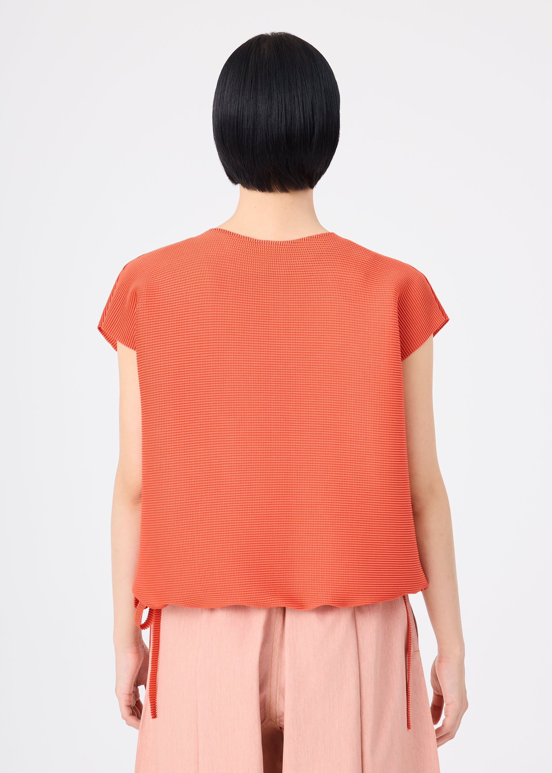 Zoomed view A person with short black hair is seen from the back against a plain light background, wearing the me ISSEY MIYAKE SQUARE STRETCH PLEATS orange round-neck top and light pink pants.