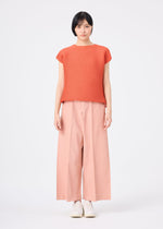 A person stands against a plain white background, wearing the me ISSEY MIYAKE SQUARE STRETCH PLEATS short-sleeved round-neck orange top, wide-legged pale pink pants, and white shoes, looking directly at the camera with a neutral expression.