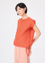 A woman with short black hair stands against a plain light background, wearing the me ISSEY MIYAKE SQUARE STRETCH PLEATS orange top and light pink wide-leg pants, looking to the side with a neutral expression.