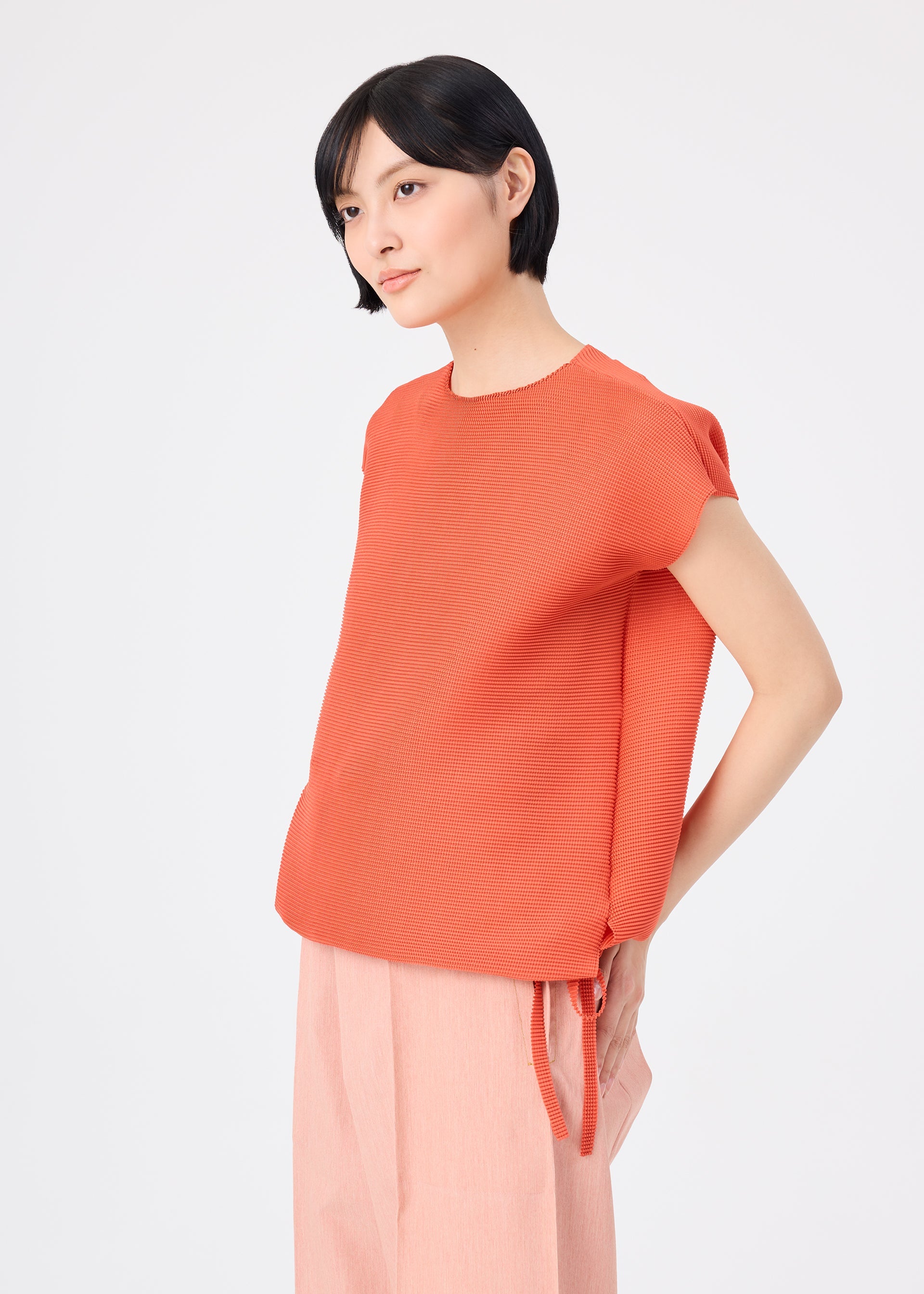 Zoomed view A woman with short black hair stands against a plain light background, wearing the me ISSEY MIYAKE SQUARE STRETCH PLEATS orange top and light pink wide-leg pants, looking to the side with a neutral expression.