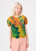 A person with light blonde hair wears the JUNGLE top by me ISSEY MIYAKE, which features bold green and orange leaf patterns, along with light pink pants, standing against a plain white background.