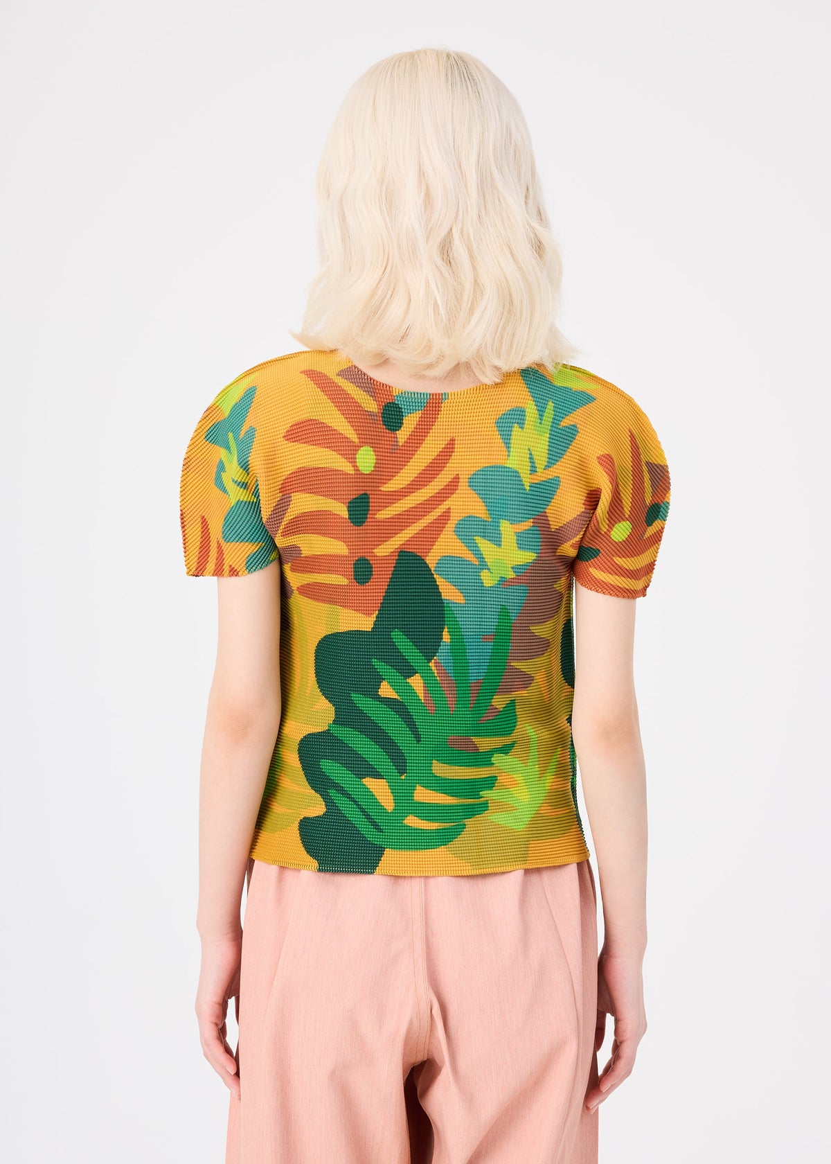 Zoomed view A person with wavy, shoulder-length blonde hair, seen from behind, wears the JUNGLE yellow pleated short sleeve shirt featuring a bold, colorful tropical leaf pattern by me ISSEY MIYAKE, paired with light pink pants. The background is plain white.