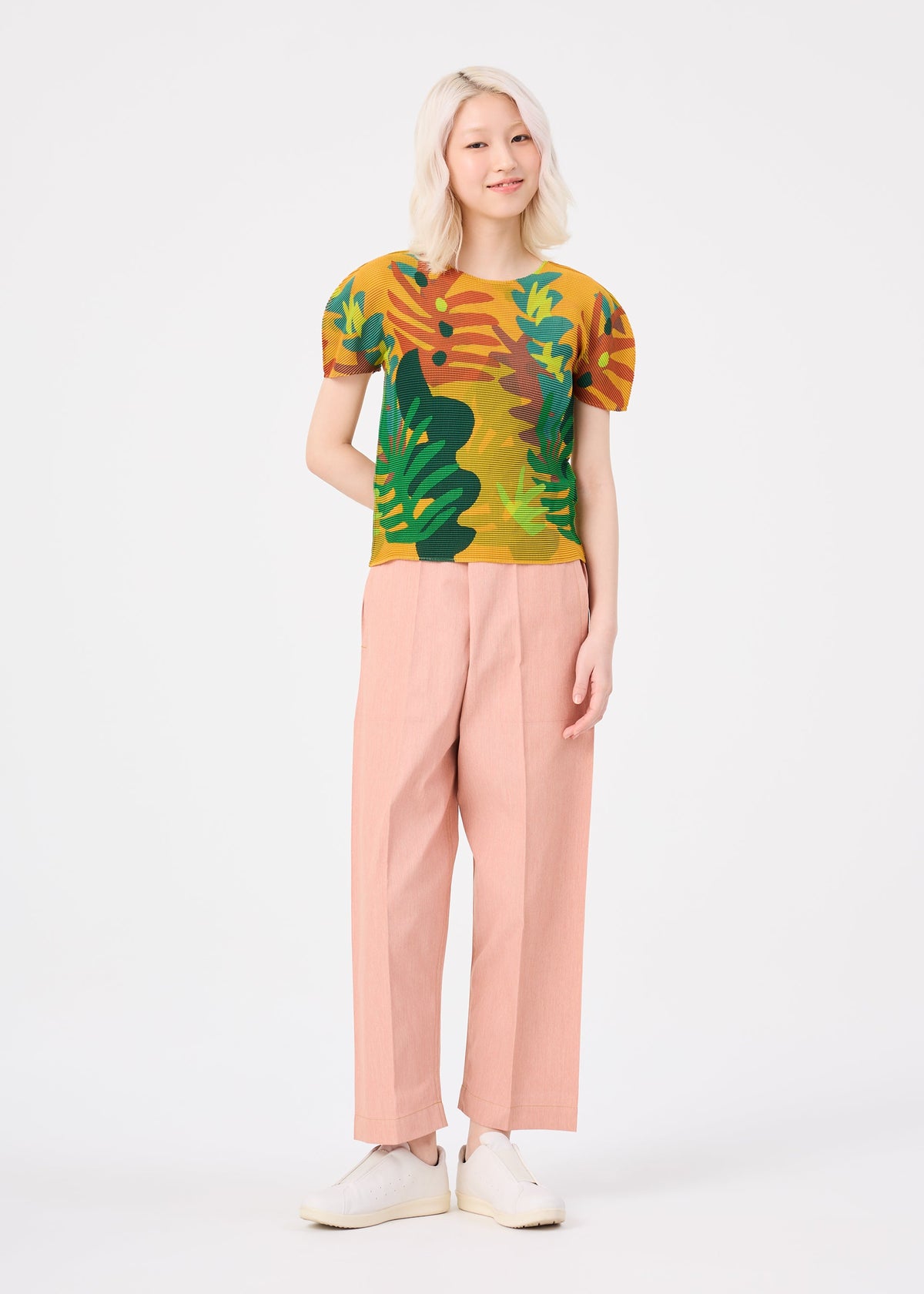 A person with light hair stands smiling against a plain white background, wearing the colorful JUNGLE top by me ISSEY MIYAKE, paired with light pink trousers and white shoes. view 6 of 6