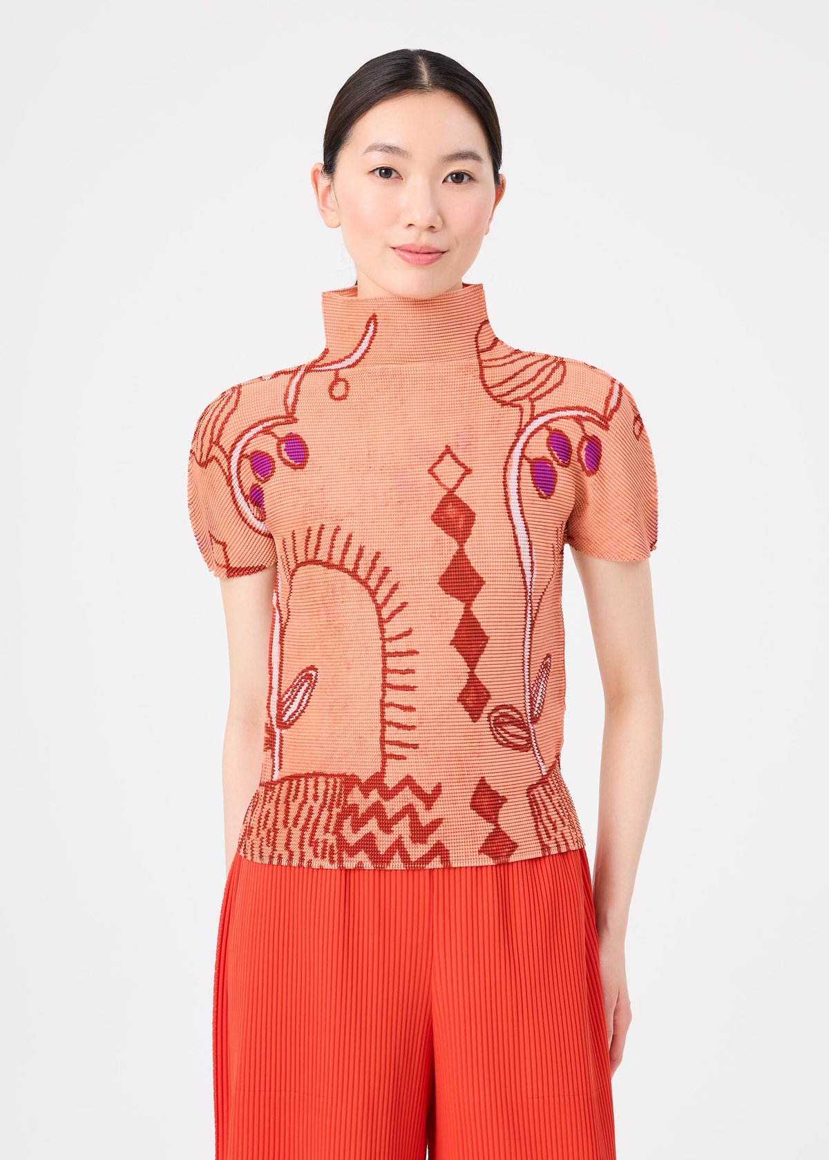 A woman stands against a plain white background wearing the “PLANT” high-neck short-sleeve top by me ISSEY MIYAKE, featuring an abstract red and purple plant pattern in peach, paired with pleated red pants. view 5 of 7