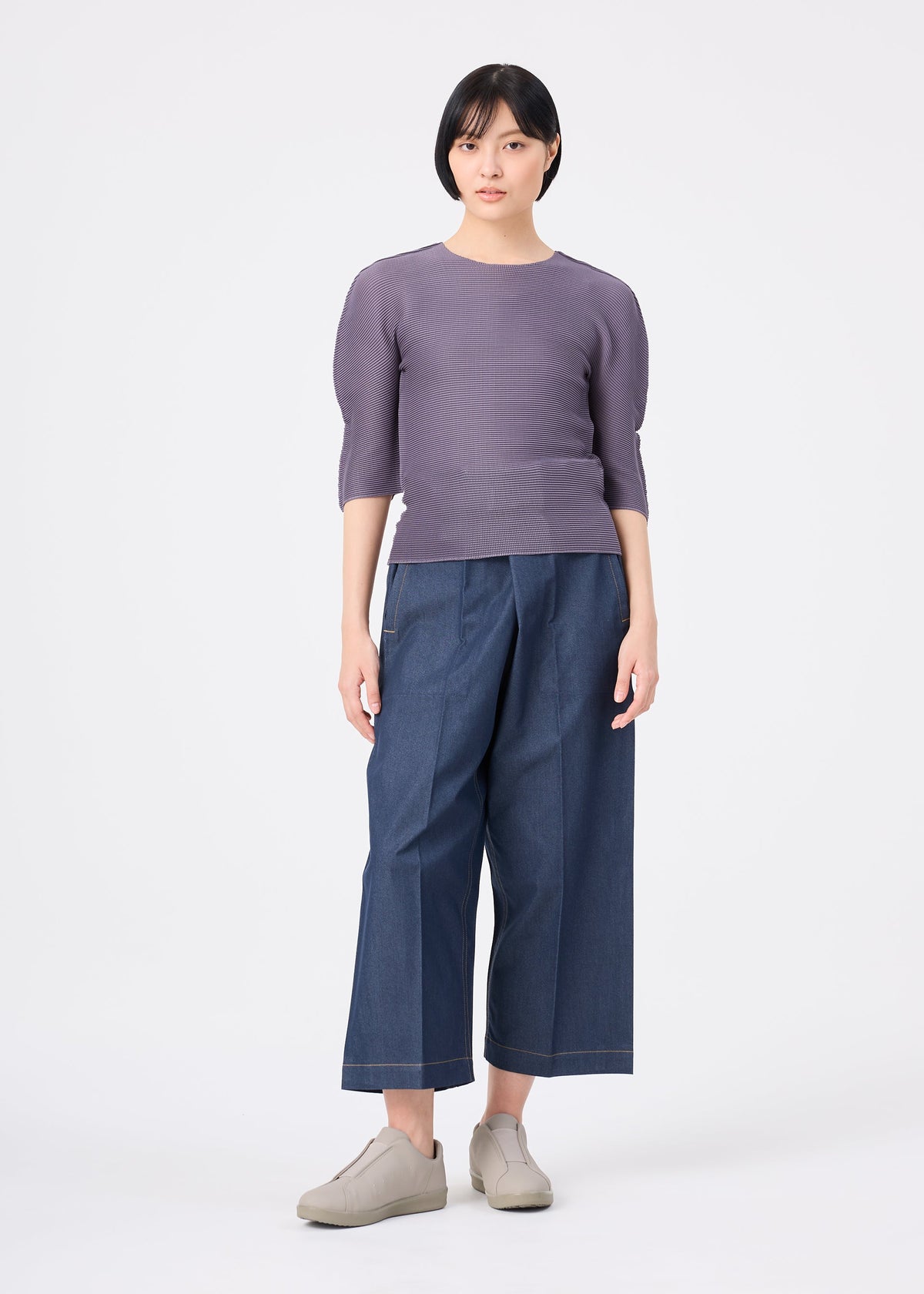 A woman with short black hair wears the STRETCH PLEATS 4 top by me ISSEY MIYAKE, paired with wide-leg blue jeans and beige shoes, standing against a plain white background. view 7 of 7