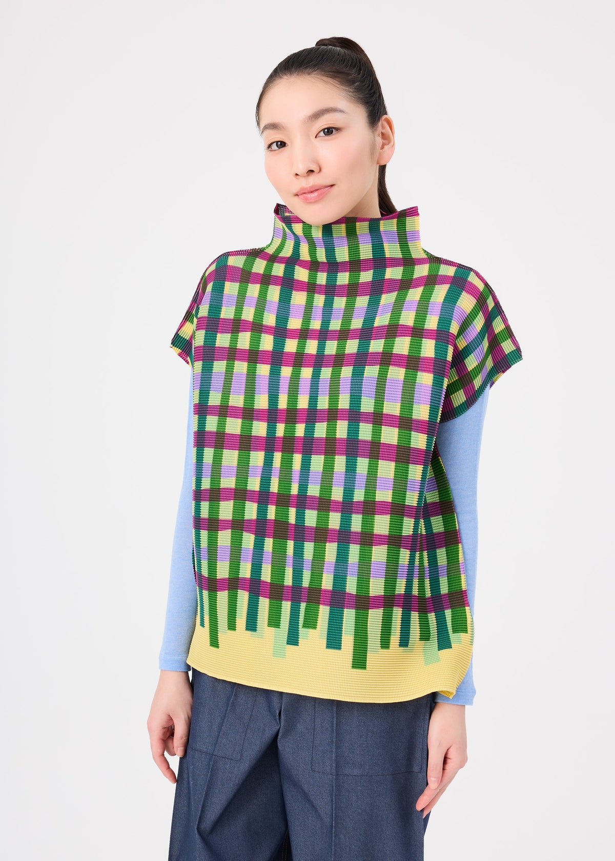 A person with long dark hair in a ponytail wears the TAPE CHECK top by me ISSEY MIYAKE over a light blue long-sleeve shirt and wide-leg pants, standing against a plain white background. view 5 of 7