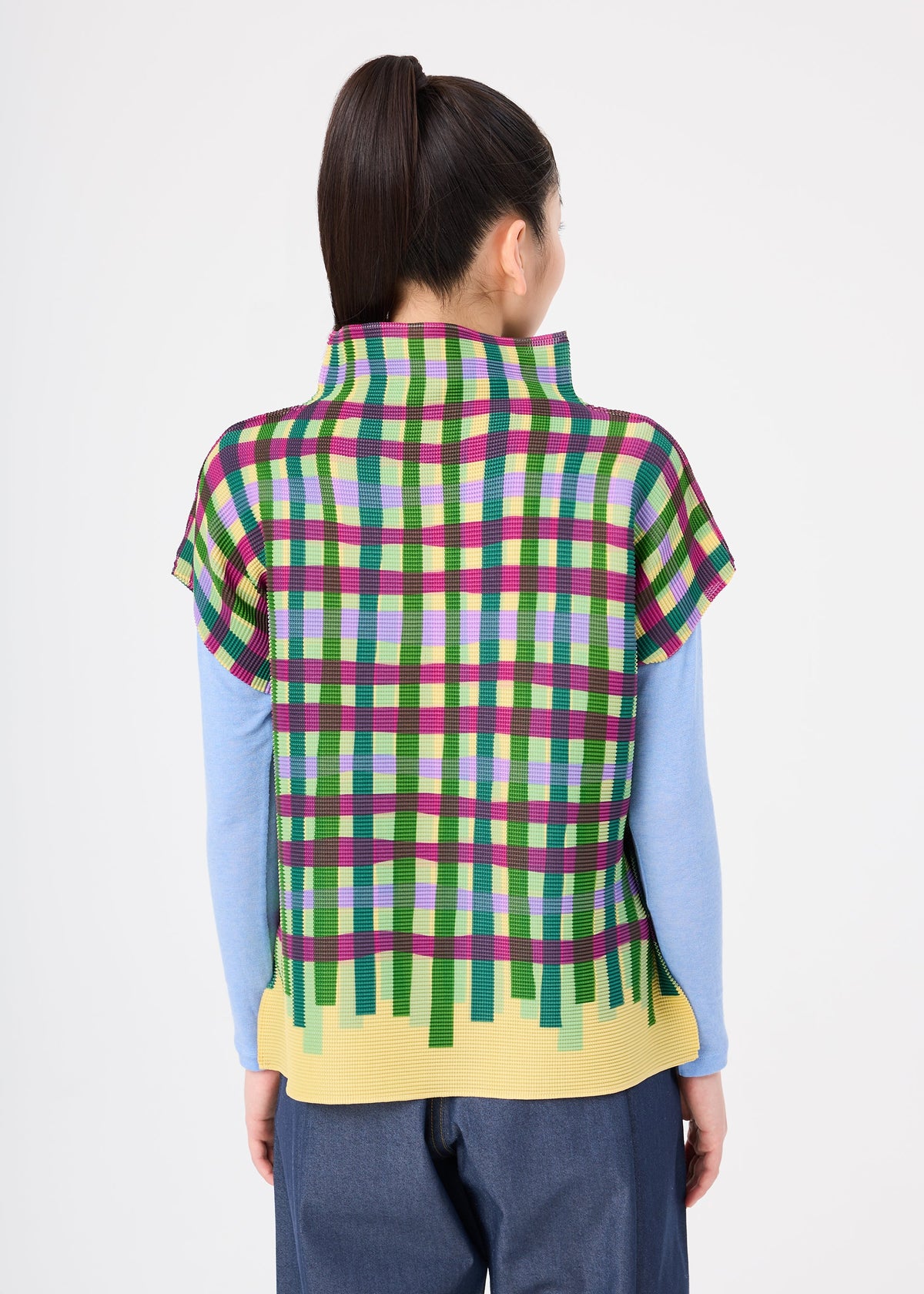 Zoomed view Person with long dark hair in a ponytail, wearing the TAPE CHECK by me ISSEY MIYAKE—a colorful plaid shirt with French sleeves and a yellow hem over a light blue high-neck top, and blue pants, facing away.
